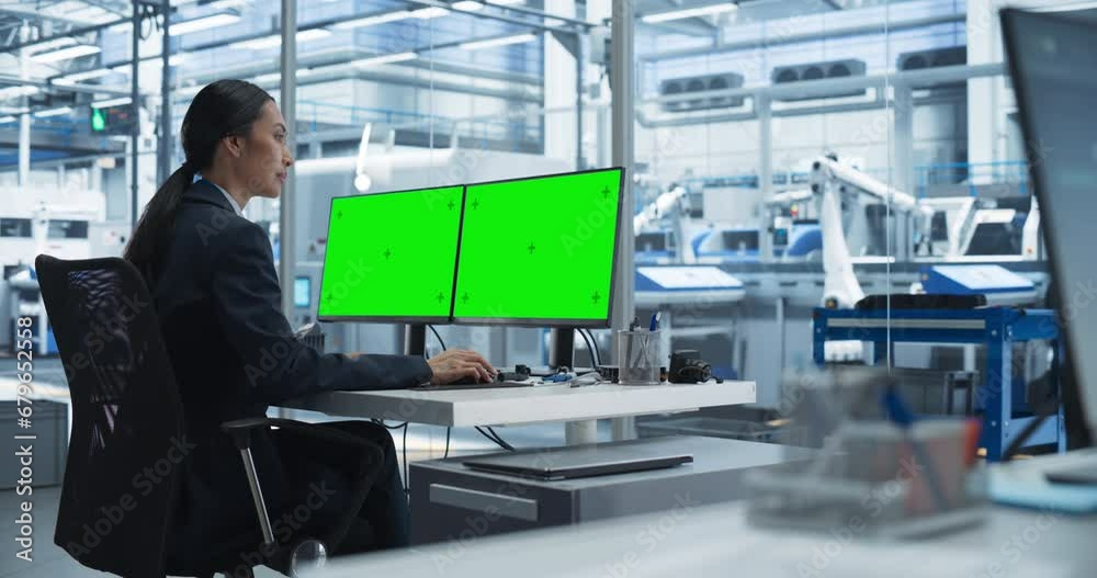 Female Robotics Technician Working on a Desktop Computer with Green Screen Chromakey Mock Up Display in a Factory Office. Technological Research and Development Facility with Autonomous Assembly Line Female Robotics Technician Working on a Desktop Computer with Green Screen Chromakey Mock Up Display in a Factory Office. Technological Research and Development Facility with Autonomous Assembly Line