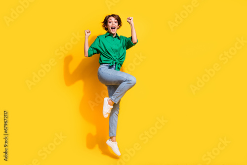 Full length photo of carefree lucky girl dressed green shirt rising fists jumping high empty space isolated yellow color background