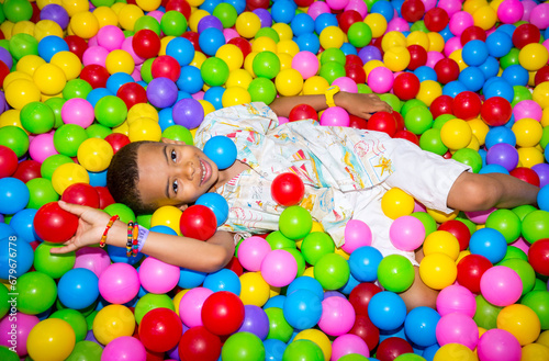 Happy african black boy playing in ball pit on birthday party in kids amusement park and indoor play center. Child playing with colorful balls in playground ball pool.