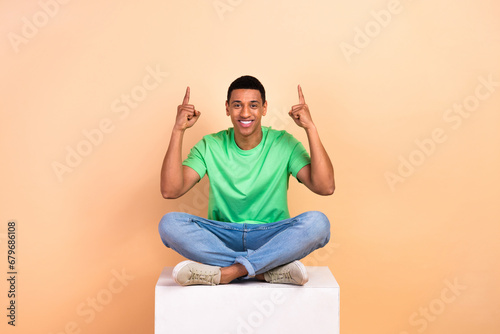 Portrait of good mood man wear stylish t-shirt sit on white cube directing at benefit empty space isolated on beige color background © deagreez