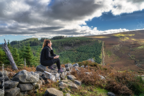 Adult woman sitting on a rock, drinking tea or coffee from thermos bottle and looking at view. Active adult, hiking in Wicklow Mountains, Ireland