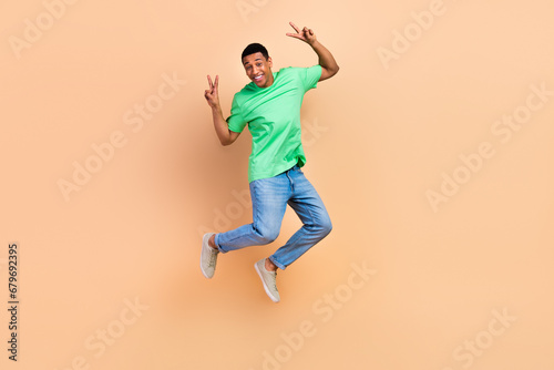 Full size photo of good mood cheerful guy dressed green t-shirt denim pants flying showing v-sign isolated on beige color background