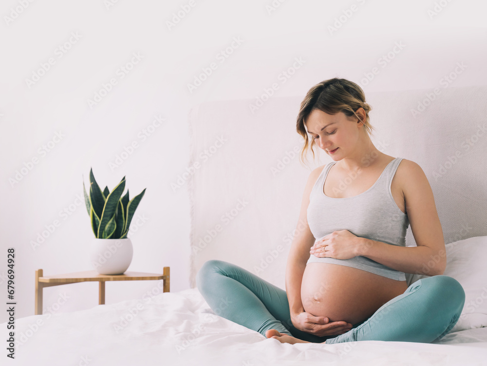 Pregnant woman in lotus pose doing meditation or breathing exercises for healthy pregnancy and preparing body for childbirth. Young expectant mother practicing yoga at home. Pregnant woman in lotus pose doing meditation or breathing exercises for healthy pregnancy and preparing body for childbirth. Young expectant mother practicing yoga at home.