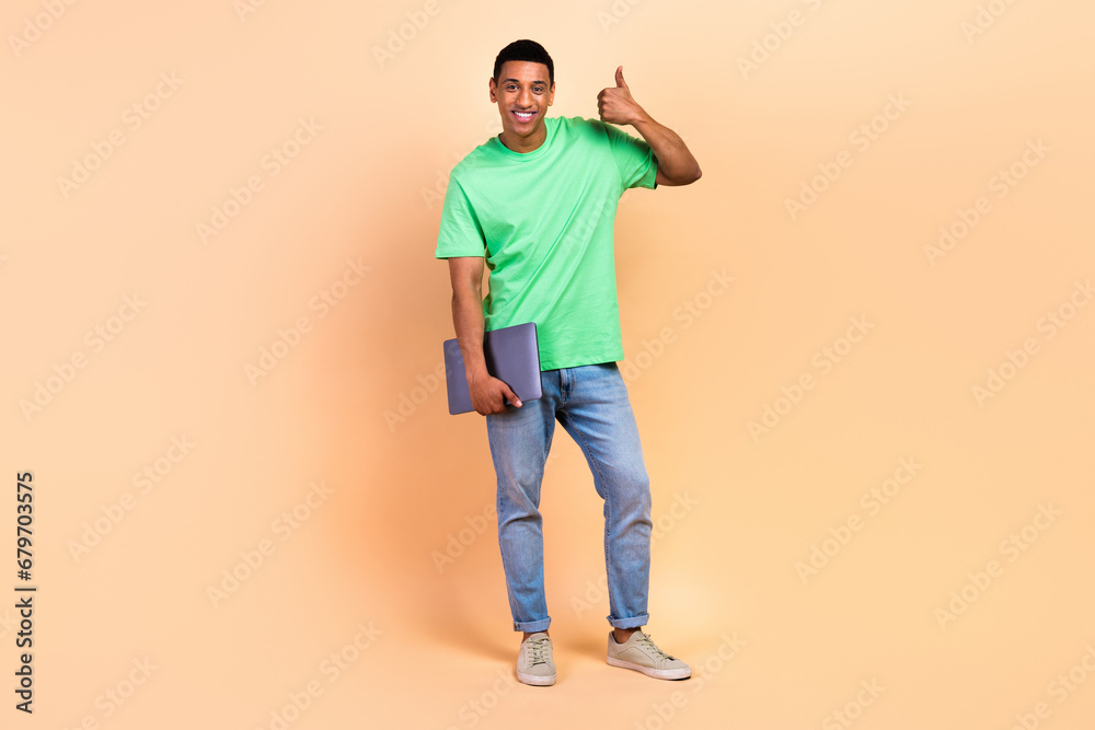 Full size photo of good mood guy dressed green t-shirt denim pants hold laptop show thumb up good job isolated on beige color background Full size photo of good mood guy dressed green t-shirt denim pants hold laptop show thumb up good job isolated on beige color background