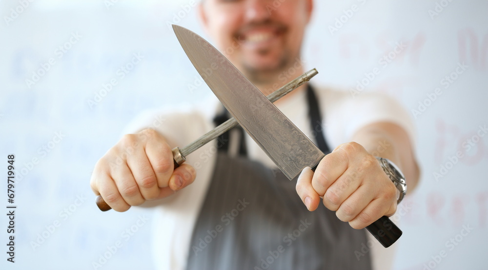 Professional Kitchen Knife Sharpening Photography. Chef Whetting Steel Blade Manually on White Background. Standing Man Holding in Hands Sharp Kitchenware for Cooking Partial View Horizontal Shot