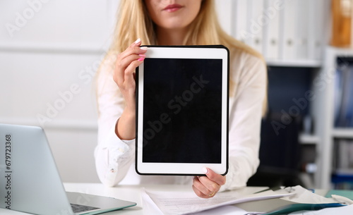 Beautiful businesswoman wearing glasses portrait at the office holding a tablet in hand sitting at the table. Smiling and looking at the camera teacher expresses success checking the test papers.