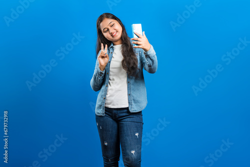 Portrait of an attractive lovely Indian girl capturing picture of oneself with victory, success gesture on mobile phone over blue backdrop