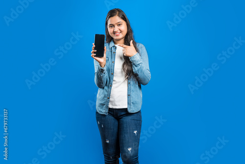 Image of a beautiful young Indian female pointing finger towards cell phone with empty screen against blue backdrop