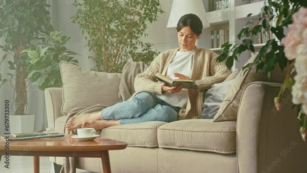 Portrait of beautiful woman relaxing, reading book while relaxing on cozy sofa at home. Smiles, enjoys leisure, carefree weekends with favorite literature and spending time. Habit, development, hobby