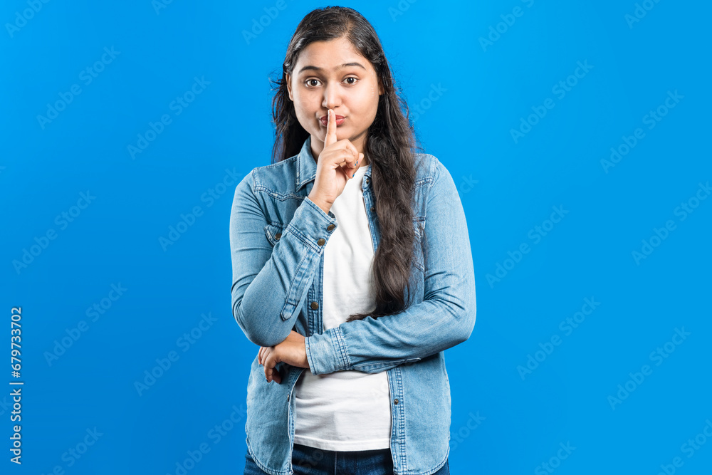 Portrait of an attractive Indian girl showing shh sign with forefinger against blue backdrop Portrait of an attractive Indian girl showing shh sign with forefinger against blue backdrop