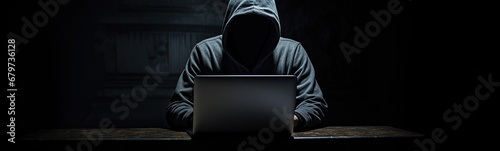 a hooded man sitting in the dark using a laptop.