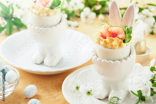Stuffed or deviled eggs with yolk, shrimp, pea microgreens with paprika in rabbit-shaped stand for easter table decorate fresh cherry or apple blossoms on light background. Traditional dish for Easter