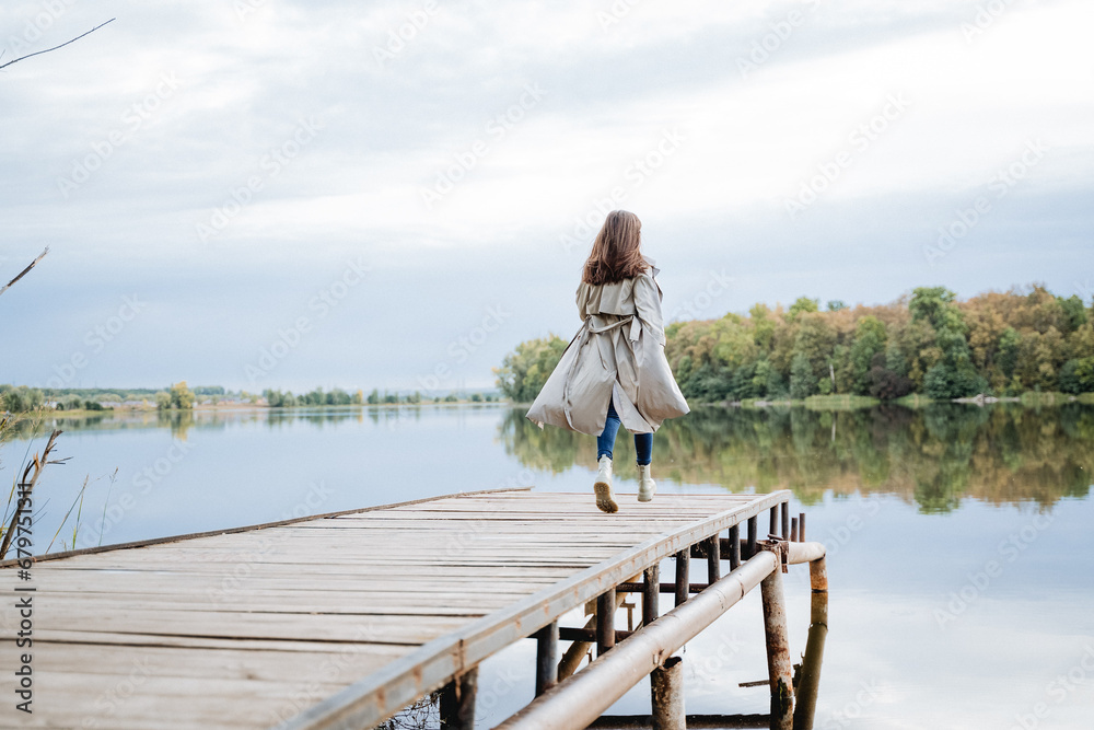 A girl in a raincoat runs along the pier on the lake. Autumn mood running in the park.