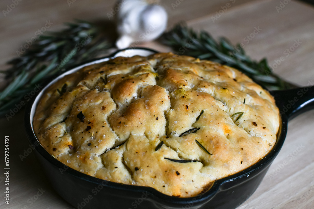 Loaf of fresh baked, homemade focaccia bread in a cast iron pan with garlic and rosemary in the background