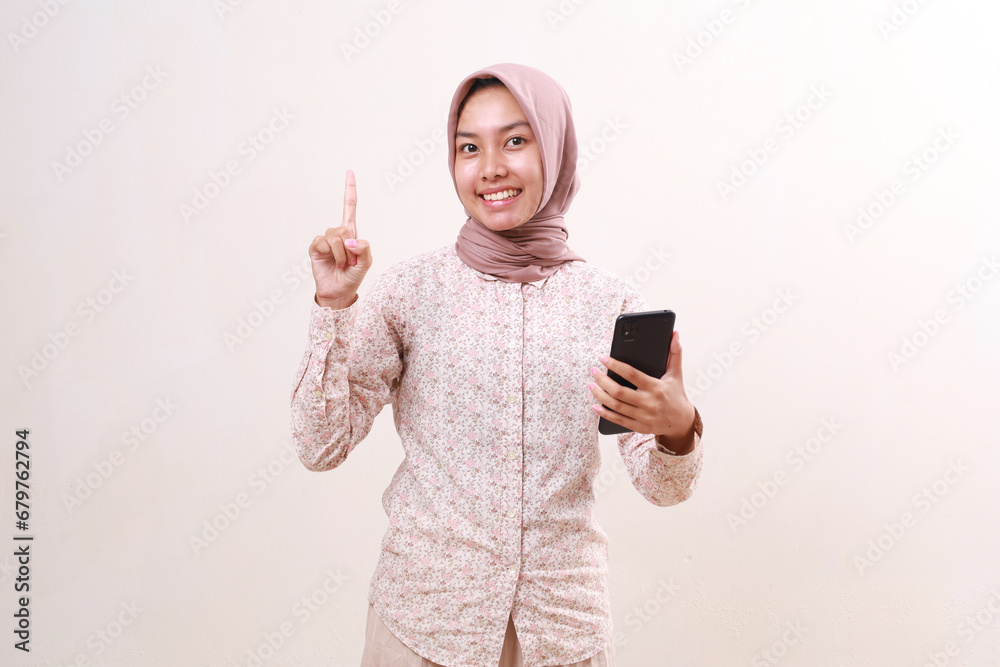 Smiling young asian muslim woman holding a cell phone while showing a finger. Isolated on white Smiling young asian muslim woman holding a cell phone while showing a finger. Isolated on white