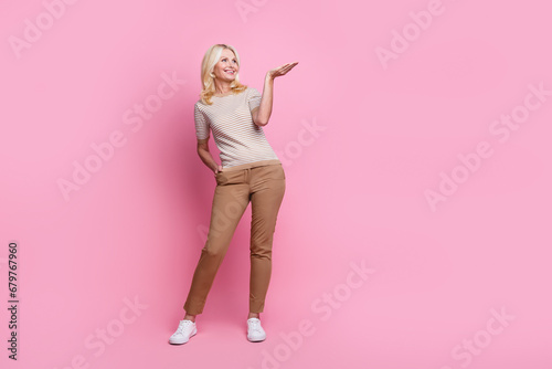 Full length photo of good mood woman wear trendy clothes brown trousers look empty space product on arm isolated on pink color background © deagreez