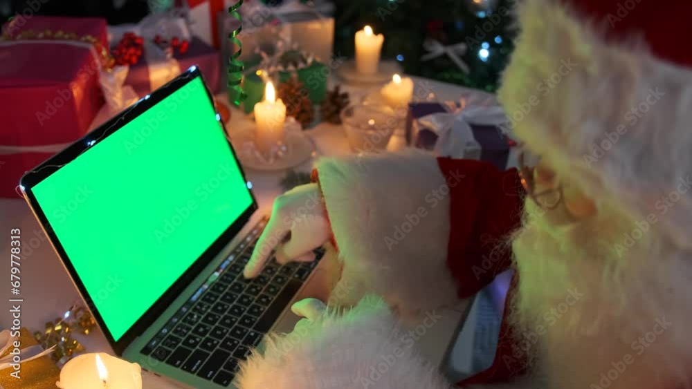 Kind Santa Claus typing on laptop with green chroma key for your AD, against the background burning candles, gifts boxes, flickering lights of Christmas tree. Happy New Year.