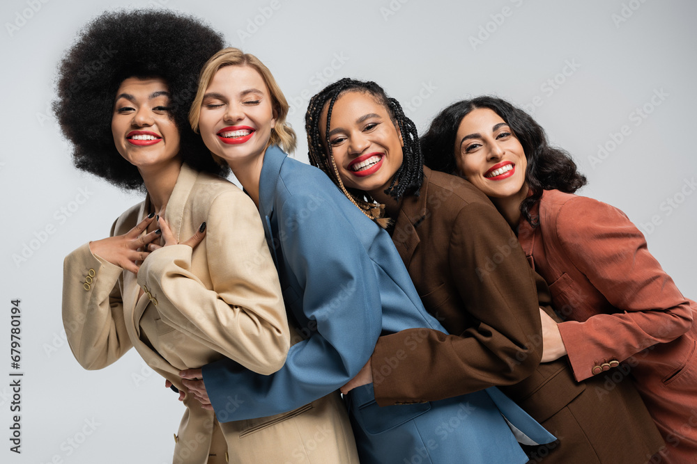 group of trendy multiracial girlfriends in colorful suits embracing and smiling at camera on grey group of trendy multiracial girlfriends in colorful suits embracing and smiling at camera on grey