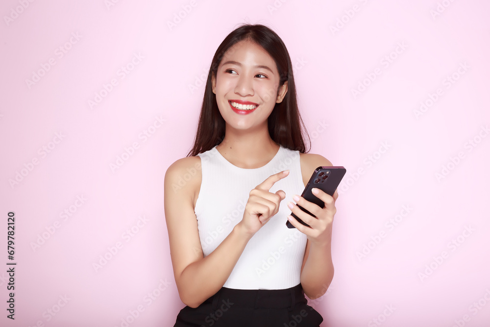 Standing and pointing happily, Portrait of a beautiful young woman in a light pink background, Asian woman holding a phone in one hand. Standing and pointing happily, Portrait of a beautiful young woman in a light pink background, Asian woman holding a phone in one hand.