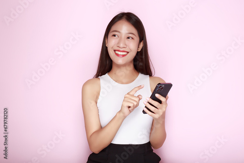 Standing and pointing happily, Portrait of a beautiful young woman in a light pink background, Asian woman holding a phone in one hand.