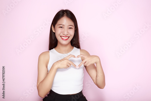 Asian woman making heart shape with hands, Portrait of a beautiful young woman in a light pink background, happy and smile, posting in stand position.