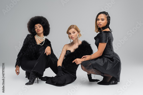 multiethnic fashionistas in black elegant wear sitting and looking at camera on grey, fashion shoot photo