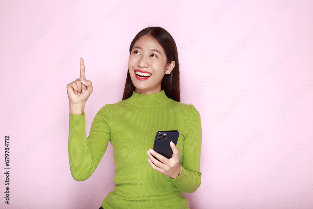 Standing and pointing happily, Portrait of a beautiful young woman in a light pink background, Asian woman holding a phone in one hand. Standing and pointing happily, Portrait of a beautiful young woman in a light pink background, Asian woman holding a phone in one hand.