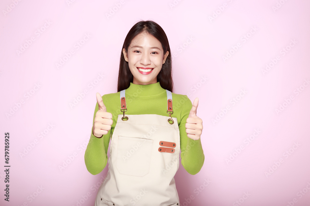 Asian woman giving two thumbs up, Portrait of a friendly young woman smiling happily, Portrait of a beautiful young woman in a light pink background, Wearing an apron. Asian woman giving two thumbs up, Portrait of a friendly young woman smiling happily, Portrait of a beautiful young woman in a light pink background, Wearing an apron.