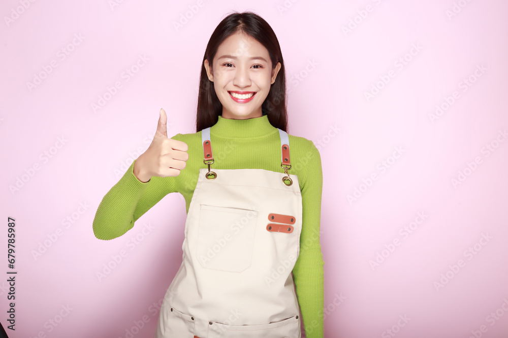Asian woman giving two thumbs up, Portrait of a friendly young woman smiling happily, Portrait of a beautiful young woman in a light pink background, Wearing an apron.