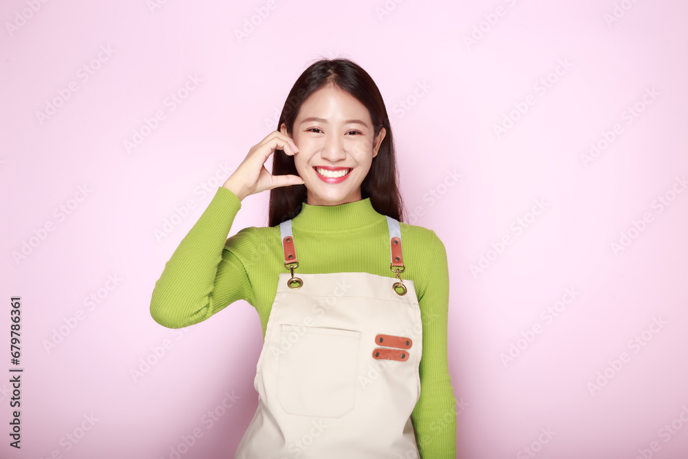Asian woman making heart shape with hands, Portrait of a beautiful young woman in a light pink background, happy and smile, posting in stand position, Wearing an apron. Asian woman making heart shape with hands, Portrait of a beautiful young woman in a light pink background, happy and smile, posting in stand position, Wearing an apron.