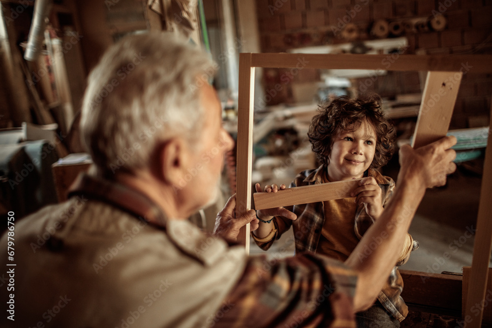 Elderly carpenter making wood frame with little boy in workshop