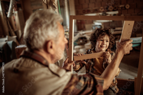 Elderly carpenter making wood frame with little boy in workshop