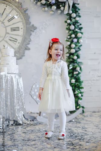 Cute little girl in a white dress with a red bandana on her head stands near the Christmas tree.