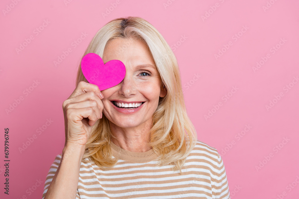 Portrait of friendly aged person toothy smile hand hold small heart symbol cover one eye empty space isolated on pink color background