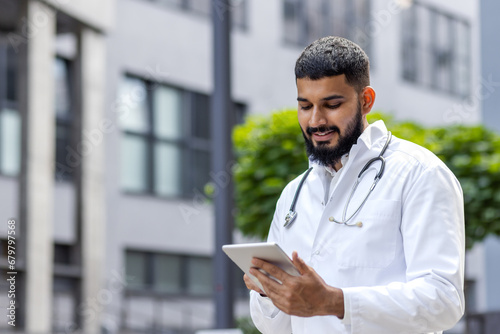A young Muslim doctor in a bedlam robe stands outside the hospital and uses a tablet, writes, types, consults