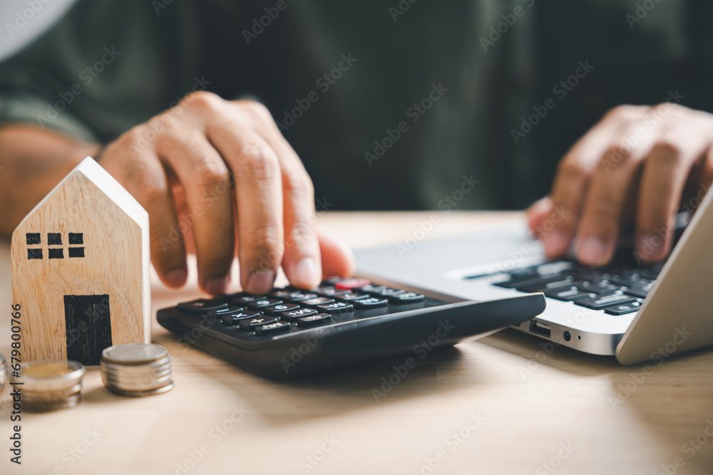 Calculators pressed by hand, aiming for home refinance. Wooden house model, buy or rent note. Concept of saving for property, strategic mortgage payment. Tax, credit analysis for financial success. - obrazy, fototapety, plakaty 