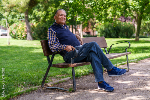 Relaxed senior man sits on a park bench, surrounded by trees, in casual attire and a contented expression