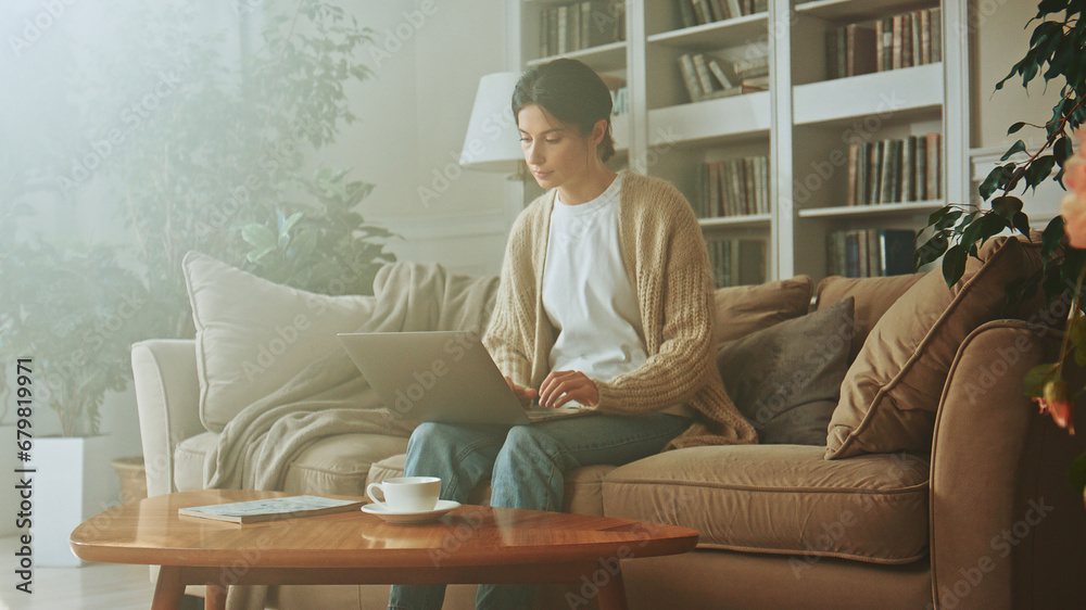 Young brunette woman uses a laptop notebook while sitting on the sofa in the living room at home. Businesswoman works remotely. Typing on a laptop keyboard while concentrating on work Young brunette woman uses a laptop notebook while sitting on the sofa in the living room at home. Businesswoman works remotely. Typing on a laptop keyboard while concentrating on work