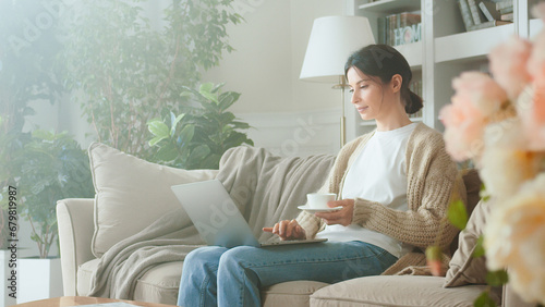 Portrait of Pretty Young Business Woman Drinking Coffee And Working Notebook From Home in Cozy Sunny Living Room. Young woman using laptop computer on sofa. Browsing on social media relaxed