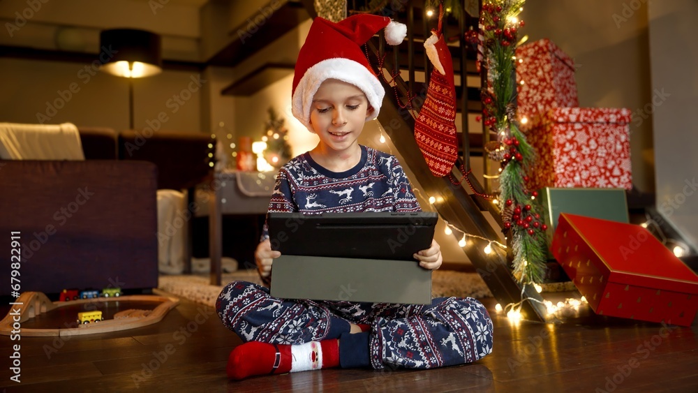 Happy smiling boy in pajamas and Santa's hat sitting on floor next to pile of gifts and presents and playing games on tablet computer. Winter holidays, celebrations and party.