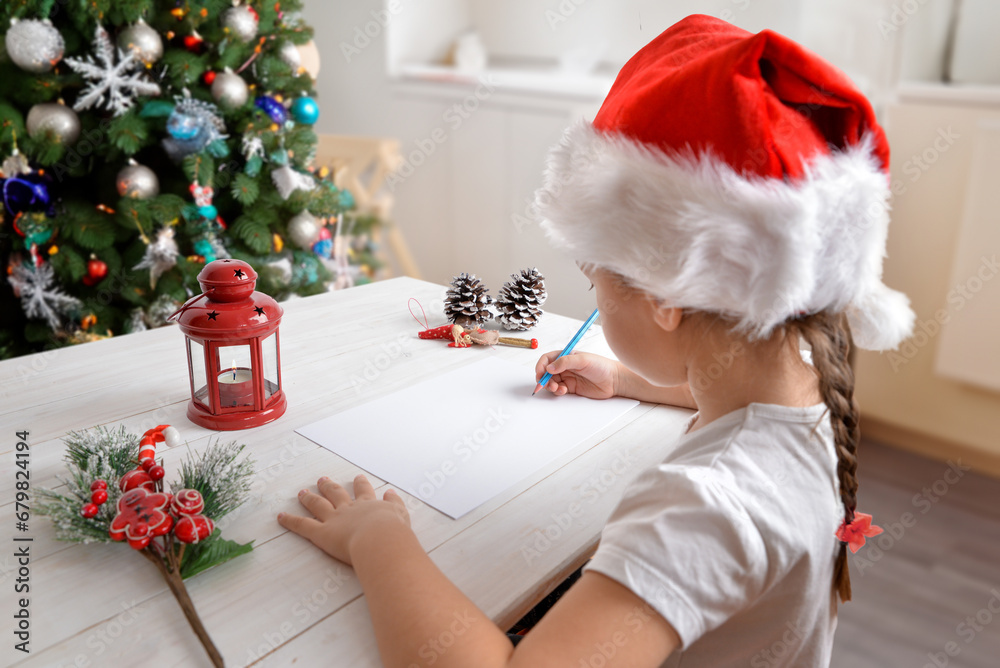 Fototapeta premium Girl writes letter to Santa Claus at home with Christmas hat. Tree and festive decorations in background. Heartwarming holiday scene