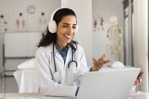 Happy beautiful young doctor woman in headphones talking to patient on video call, using laptop for online conference chat, giving professional consultation on Internet from hospital workplace photo