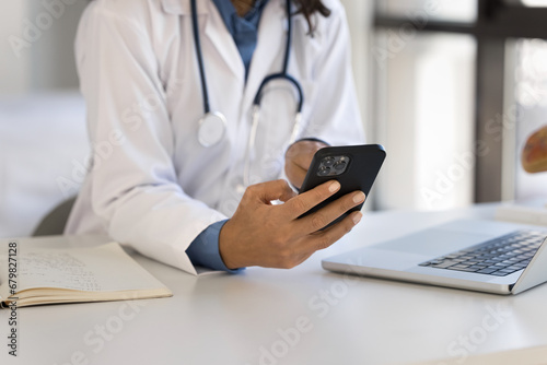 Hands of young female doctor using smartphone at workplace, sitting at laptop, working with medical professional app, chatting with patient, giving consultation on Internet