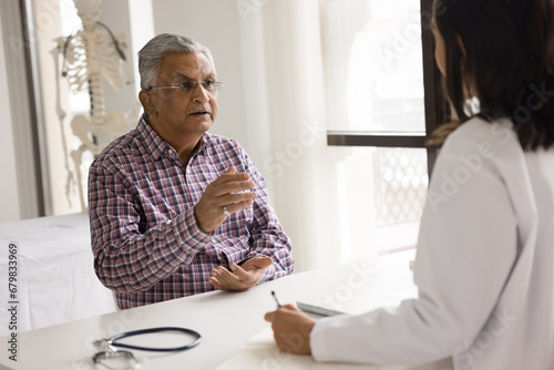 Serious elderly Indian man visiting younger doctor woman for healthcare examination, speaking, complaining on symptoms, consulting practitioner about therapy, treatment, checkup