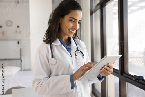 Positive young Latin practitioner woman working in clinic office with digital tablet computer, giving online consultation, checking electronic medical records in hospital application