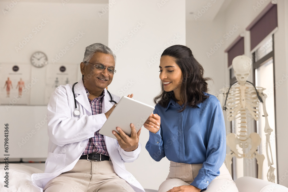 Fototapeta premium Positive multiethnic mature doctor man and younger patient woman using online medical application on digital tablet together, smiling, laughing, discussing examination results, treatment