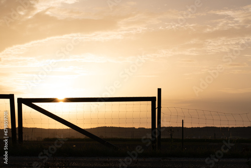 The sun sets behind the horizon. An old fence made of wood and wire stands at the side of the road. Fields can be seen.