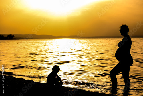 Silhouette of pregnant mother and child, sunset over sea. Family summer vacation concept.