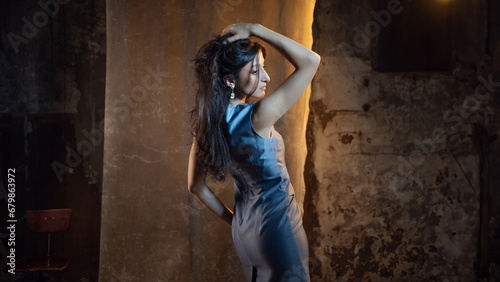 Elegant and beautiful brunette in a chic evening dress, a blue dress of medium length. photo in dark colors on a textured wall, warm light