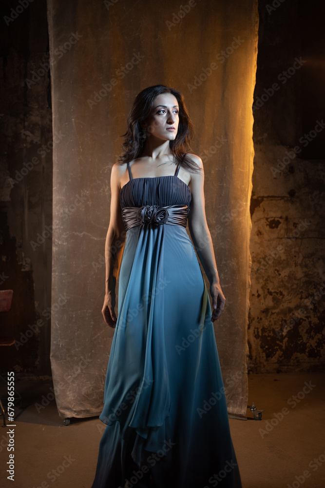 An elegant and beautiful brunette in a chic evening dress, a blue dress with a long flowing skirt to the floor. photo in dark colors on a textured wall, warm light